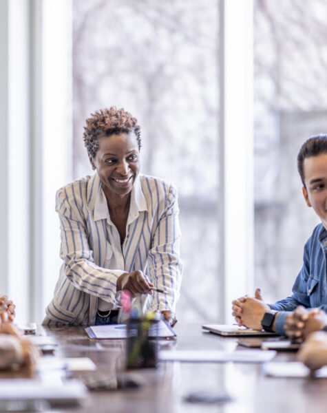 Brainstorming in a Business Meeting A small group of business professionals sit around a table as they meet to brainstorm some ideas for the future of the company. They are each dressed professionally and have papers scattered out in front of them as they work together. The focus is on a woman of African decent who is pitching an idea to the group.