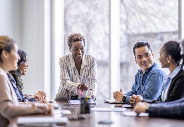 A small group of business professionals sit around a table as they meet to brainstorm some ideas for the future of the company. They are each dressed professionally and have papers scattered out in front of them as they work together. The focus is on a woman of African decent who is pitching an idea to the group.